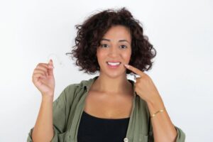 Woman holding Invisalign aligner and pointing at her smile 